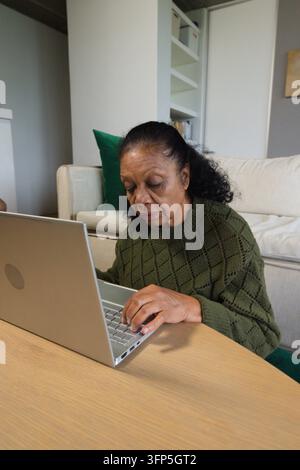 Donna anziana afroamericana che digita su un laptop argentato a un tavolo basso a casa, indossando un maglione verde Foto Stock