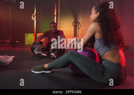 Amici diversi che si riposano e chiacchierano in abbigliamento sportivo su un tappeto da ginnastica con palle mediche viola e nere. Palestra, fitness, allenamento, forza, anelli, pesi, Foto Stock