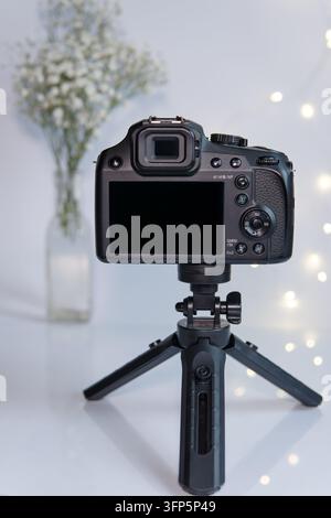 Fotocamera digitale nera su mini treppiede con schermo vuoto e sfondo luminoso floreale accogliente – modello ideale per corsi di fotografia, creatori di contenuti, blog Foto Stock