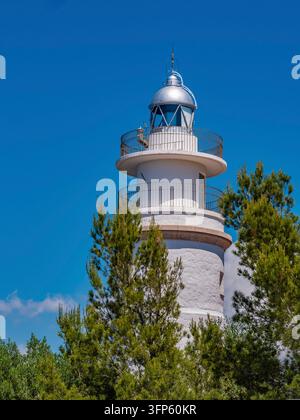 Torre cilindrica bianca di faro si erge contro il brillante cielo blu mediterraneo a Port de Soller, incorniciata da lussureggianti pini verdi - costa classica Foto Stock