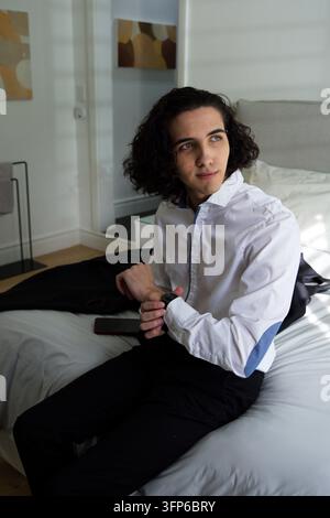 Uomo afroamericano sulla ventina che aggiusta l'orologio mentre è seduto in camera da letto con una camicia bianca e il telefono Foto Stock