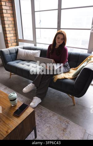 Donna seduta su un divano grigio trapuntato in un loft, utilizza un laptop con smartphone e tazza da viaggio nelle vicinanze Foto Stock