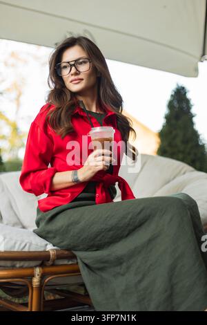 Giovane donna elegante con occhiali e camicetta rossa seduta su una comoda poltrona nella terrazza esterna di un caffè, con in mano una tazza di plastica di caffè freddo Foto Stock