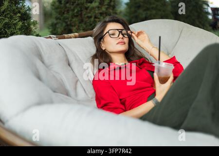 Giovane donna elegante con occhiali e una camicia rossa che aggiusta la sua montatura mentre tiene un caffè freddo, sdraiata con sicurezza su un comodo divano imbottito all'aperto Foto Stock