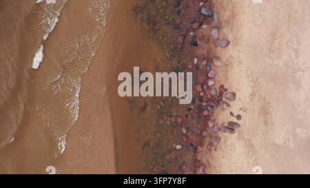 Vista aerea di onde che si infrangono su una spiaggia sabbiosa con ciottoli colorati sparsi lungo la riva a Latvija Foto Stock
