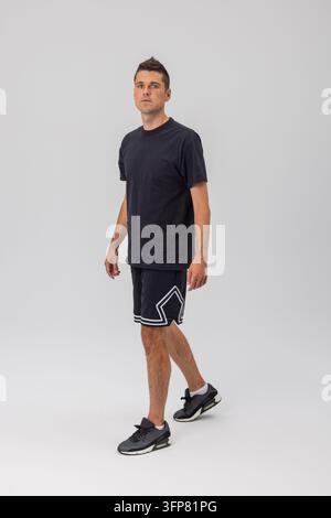 Un giovane uomo atletico cammina in un ambiente da studio, indossa una maglietta nera semplice e pantaloncini sportivi a fantasia, guardando avanti. Foto a figura intera contro un Foto Stock