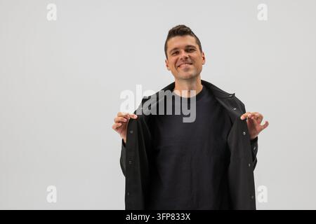 Uomo felice e affascinante che sorride alla telecamera tenendo aperta la sua giacca nera, isolato su sfondo grigio chiaro in studio. Ritratto maschile allegro per la moda, appa Foto Stock