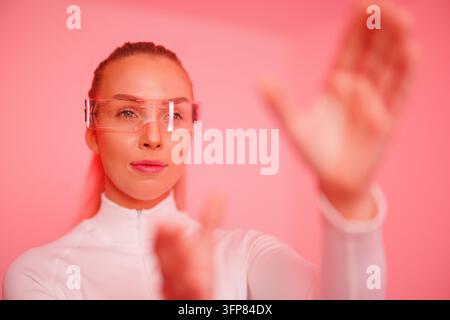 Giovane donna sicura di sé con smartglasses che gesticola su uno sfondo neon rosso Foto Stock