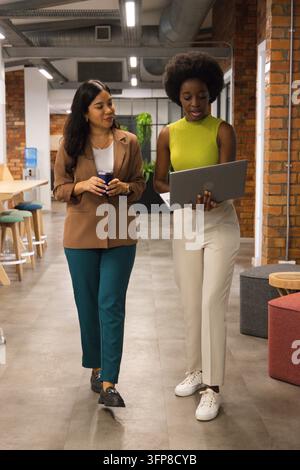 Diverse colleghe donne che camminano fianco a fianco lungo un moderno corridoio d'ufficio tenendo un laptop aperto e una tazza Foto Stock