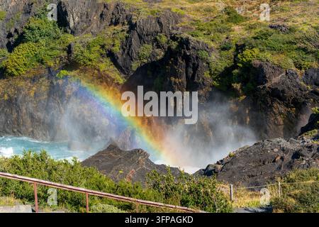 Vista delle cascate Salto Chico con un arcobaleno vivace sopra l'acqua che cade a cascata, circondata da rocce scure e frastagliate e vegetazione verde a Torres del Paine, M Foto Stock