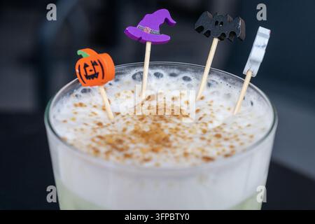 un latte freddo schiumoso ricoperto di schiuma alla cannella spolverata e decorato con divertenti decorazioni di stuzzicadenti di Halloween tra cui una zucca, un cappello da strega, un pipistrello e g Foto Stock