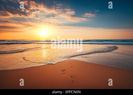 Impronte sulla spiaggia sabbiosa che conducono all'oceano al tramonto Foto Stock