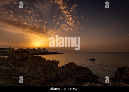 Tramonto dorato su un mare calmo con scogli costieri frastagliati, una piccola barca da pesca e edifici lontani sul lungomare sotto nuvole morbide. Foto Stock