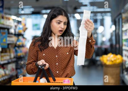 Donna che tiene un lungo scontrino della spesa ed esprime shock e incredulità mentre è in piedi in un corridoio del supermercato, riflettendo l'aumento dei costi e le preoccupazioni sul budget Foto Stock