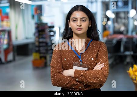 Giovane donna professionista con un badge identificativo e le braccia incrociate, in piedi seriamente in un corridoio di un negozio di alimentari o supermercato, rappresentando l'occupazione nella vendita al dettaglio e il servizio clienti Foto Stock