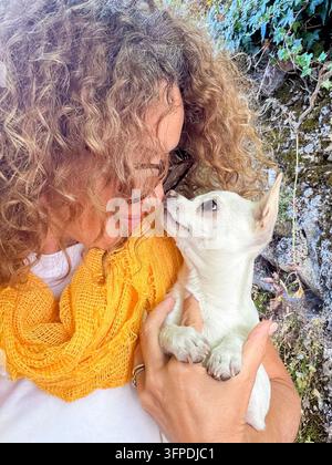 Donna con capelli ricci e occhiali che tiene vicino un piccolo chihuahua bianco, condividendo un affettuoso tocco di naso mentre indossa una sciarpa gialla brillante all'aperto contro un muro di pietra con edera. Foto Stock