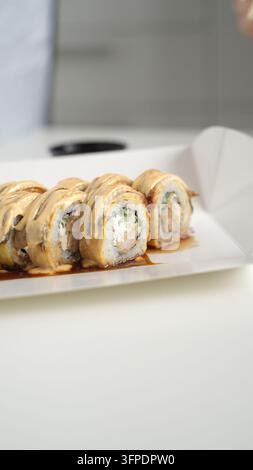Rotoli di sushi guarniti con salsa cremosa e anguilla grigliata, serviti in una scatola da asporto bianca per un pasto asiatico comodo e gustoso Foto Stock