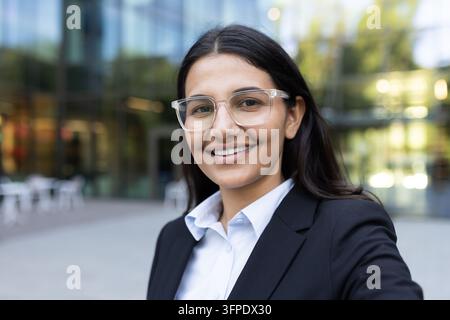 Giovane donna professionista con occhiali e abito, che sorride direttamente alla fotocamera mentre è in piedi davanti a un edificio moderno di uffici, simboleggiando successo e leadership Foto Stock