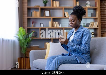 Giovane donna nera seduta comodamente su un divano in un soggiorno, sorridendo mentre interagisce con il suo smartphone, godendosi il tempo libero e la connettività digitale a casa Foto Stock