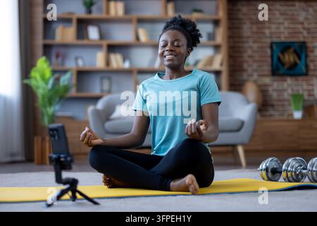 Giovane donna afroamericana che pratica yoga e meditazione in posizione del loto su un tappetino, registrando un video con lo smartphone per il suo corso di fitness online a casa Foto Stock