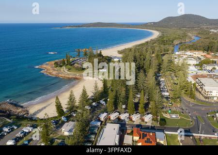 Veduta aerea di Horseshoe Beach accanto a un campeggio ombreggiato da alti alberi, con Front Beach che si snoda attorno alla baia sullo sfondo. Promontorio roccioso Foto Stock