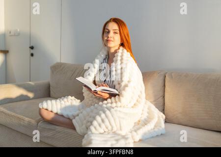 Donna che legge un libro avvolta in una coperta accogliente su un divano alla luce del mattino. Foto Stock