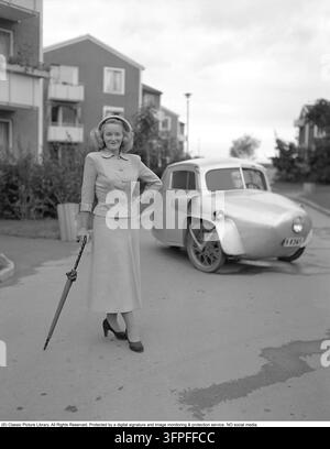Lusso quotidiano elegante nel 1949 – un'epoca in cui la moda femminile era definita da silhouette aderenti, vita accentuata e semplicità sofisticata. La giacca e la gonna abbinate riflettono l'estetica del dopoguerra – un abito tipico per la donna svedese moderna che desiderava essere sia pratica che chic. Questa silhouette a clessidra è stata ispirata dal rivoluzionario "New Look" di Christian Dior, che ha sconvolto il mondo della moda nel 1947. Dietro di lei si trova una microauto a tre ruote, una costruzione personalizzata e fatta in casa che utilizza ruote da moto nella parte anteriore. Nell'Europa del dopoguerra, gravi carenze di materiale Foto Stock