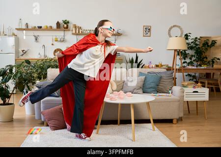 Una ragazza allegra con un mantello da supereroe rosso gioca felicemente nella sua accogliente casa Foto Stock