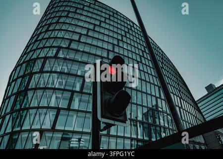 Grattacielo moderno in vetro che si incurva verso l'alto, con un semaforo rosso e architettura urbana contro un cielo blu-verde Foto Stock