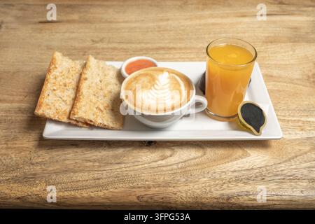Combo colazione con pane tostato, un cappuccino con latte art, succo d'arancia e una piccola ciotola di marmellata su un piatto bianco rettangolare. Foto Stock