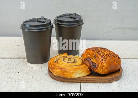 Due tazze di caffè da asporto nere con coperchio accanto a una pasta a spirale e un panino glassato su un piccolo vassoio di legno. Foto Stock