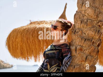 Donna con occhiali da sole e vestito stampato appoggiata al tronco di una palma in un resort sulla spiaggia con ombrelloni di paglia e cielo blu sullo sfondo. Foto Stock