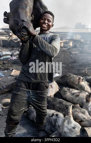 Un giovane uomo sorridente porta una testa di bestiame bruciacchiata sulla spalla in un fumoso mercato del bestiame sulla spiaggia, Regione di Greater Accra, Accra, Ghana Foto Stock