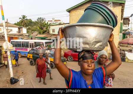 Donna che bilancia una pila di bacinelle di plastica sulla testa mentre cammina in un mercato affollato, Regione Centrale, Elmina, Ghana Foto Stock