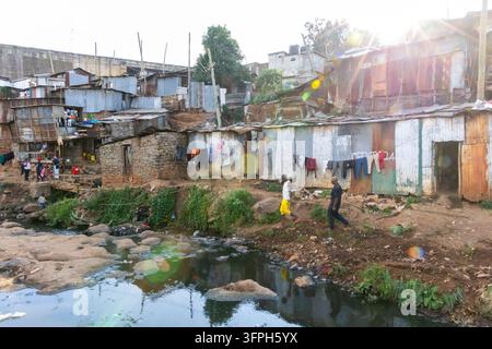 Due uomini che camminano lungo un torrente inquinato accanto a baracche di lamiera ondulata e lavanderia nell'area di Kibera, Contea di Nairobi, Nairobi, Kenya Foto Stock