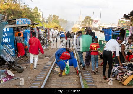 Donna seduta su una tanica sulla linea ferroviaria tra bancarelle del mercato e persone che camminano nell'area di Kibera, Contea di Nairobi, Nairobi, Kenya Foto Stock