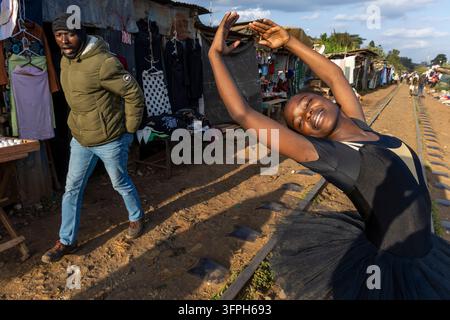 Ballerina sorridente in tutù nero che si inarca con le braccia alzate sui binari ferroviari della scuola di balletto di Kibera mentre un uomo con una giacca verde passa oltre Foto Stock