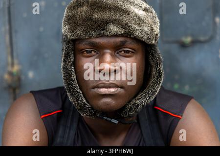 Ritratto ravvicinato di un giovane uomo nell'area di Kibera che indossa un cappello di finta pelliccia grigio e una maglietta nera senza maniche davanti a un muro blu grigio, Contea di Nairobi, Nai Foto Stock
