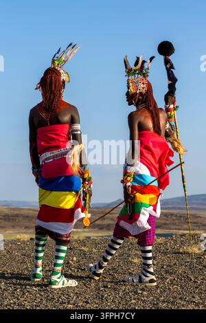 Due uomini della tribù Rendille visti di spalle con abiti colorati e copricapi piumati che tengono un'asta di bandiera e una lancia su terreno roccioso, Contea di Marsabit, L Foto Stock