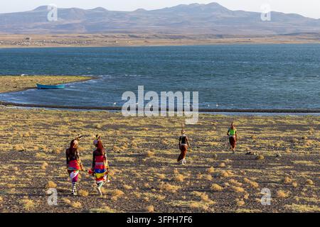 Quattro uomini e donne della tribù Rendille in costumi colorati che camminano su una riva erbosa verso il lago Turkana dove i fenicotteri stanno nelle acque basse, Ma Foto Stock