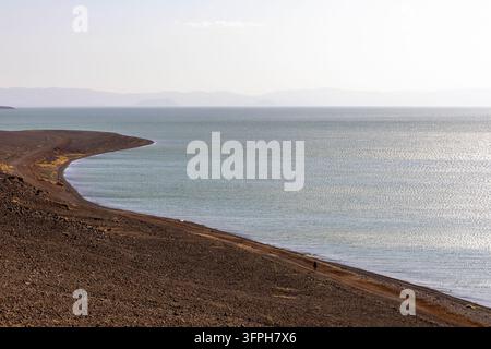 Piccola figura solitaria che cammina lungo una riva di ghiaia curva di un vasto lago calmo con montagne sfocate, Contea di Marsabit, Loiyangalani, Kenya Foto Stock