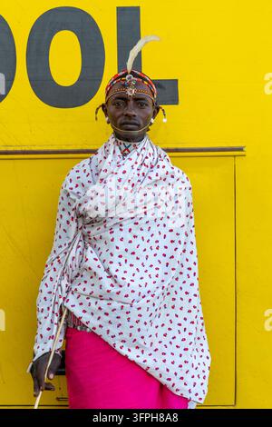 Uomo della tribù Rendille con una fascia per capelli decorata con perline e una piuma e uno scialle floreale bianco, in piedi davanti a un muro giallo brillante mentre tiene un bastone sottile, Marsa Foto Stock