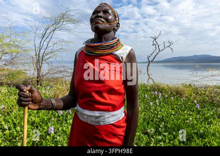 Donna della tribù Njemps in un vestito rosso e collane di perle che guarda verso l'alto mentre tiene un bastone accanto al lago con giacinti d'acqua, Lago Baringo, Kenya Foto Stock