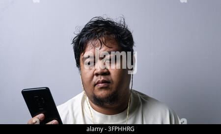Un ritratto frontale di un uomo asiatico adulto che guarda in basso uno smartphone nero tenuto in mano. Questo soggetto indossa una maglietta a maniche lunghe color crema casual e wir Foto Stock