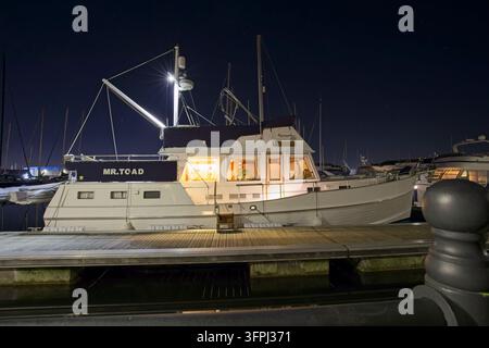 Vista laterale di uno yacht di lusso bianco chiamato "Mr. Toad" con luci calde nella cabina interna, ormeggiato lungo un pontile in legno di notte. Foto Stock