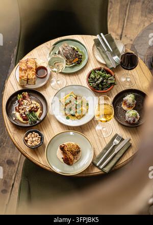 Vista aerea di un tavolo rotondo in legno disposto con diversi piccoli piatti gourmet, pesce arrosto, asparagi avvolti nel prosciutto e bicchiere di vino pregiato Foto Stock