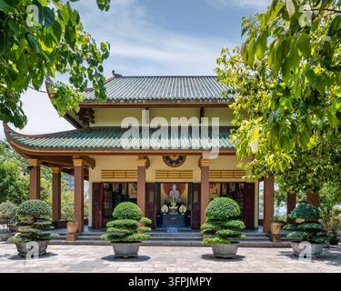Architettura tradizionale del tempio con una statua di Buddha al pagoda Linh Ung a Da Nang, Vietnam, incorniciata da alberi verdi e piante bonsai decorative Foto Stock