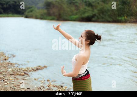 Una giovane donna asiatica che pratica la meditazione accanto a un fiume calmo, alzando le braccia con concentrazione pacifica vicino a una riva rocciosa e una foresta verde Foto Stock