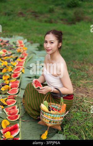 Una donna asiatica che tiene una anguria accanto a una esposizione di frutta colorata in un ambiente erboso all'aperto Foto Stock