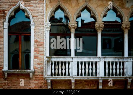 Vista ravvicinata di una vecchia facciata di palazzo veneziano con archi gotici appuntiti, colonne di marmo, balcone in pietra e finestre scure riflettenti su mura consumate dal tempo. Foto Stock